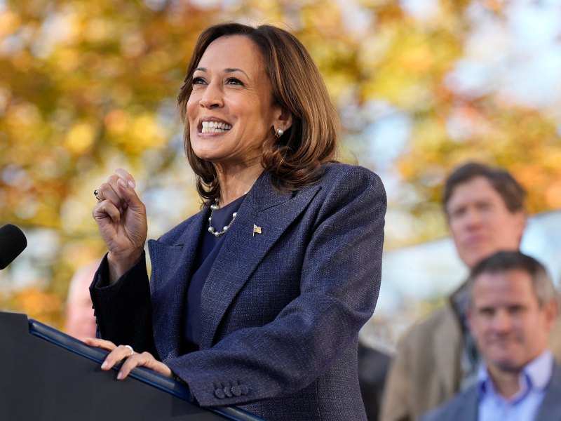 A woman speaks at a podium outside, the leaves in the trees have autumn colors