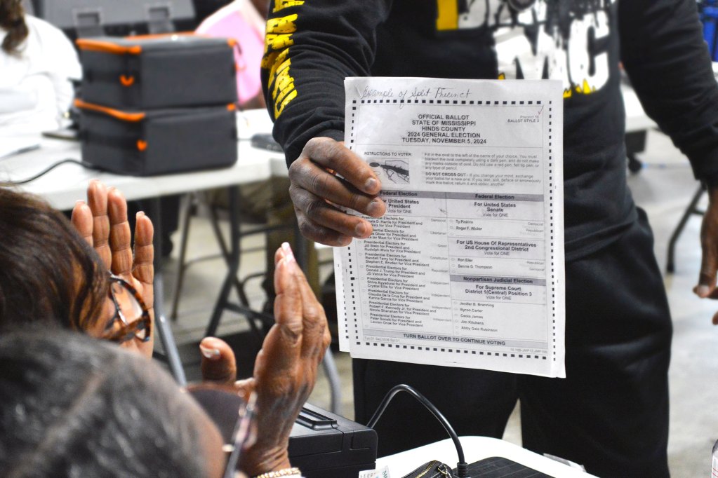A man holds up an example of a voting ballot before a woman