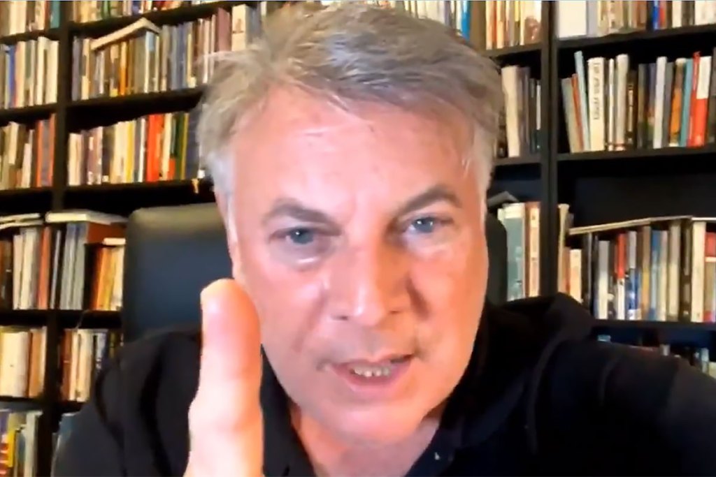 A man sits in front of a bookshelf and speaks and points with his hand
