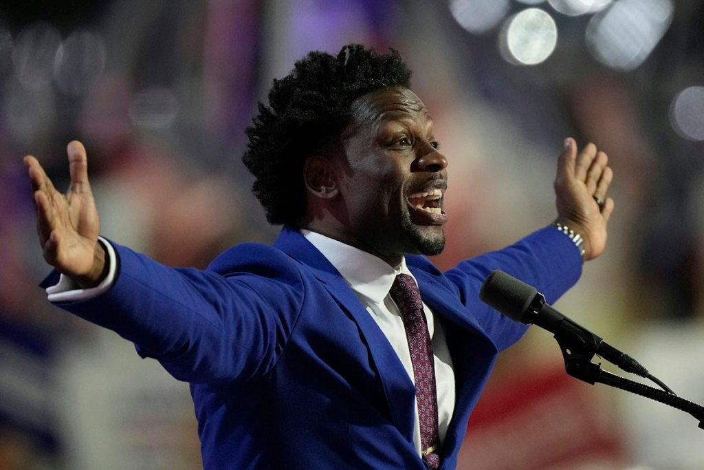 A Black man in a blue suit holds out his arms as he speaks into a microphone.