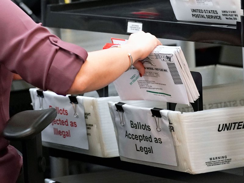 A hand is visibly moving mail in ballots to a box marked "Ballots accepted as legal"