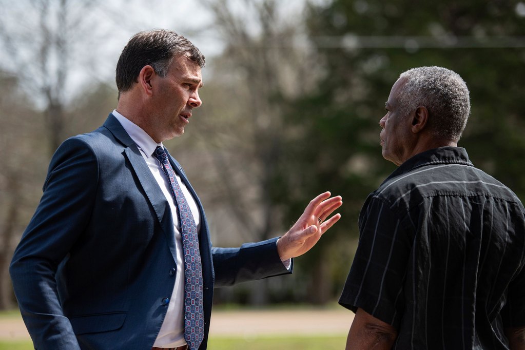 A man in a suit speaks to another man to his right