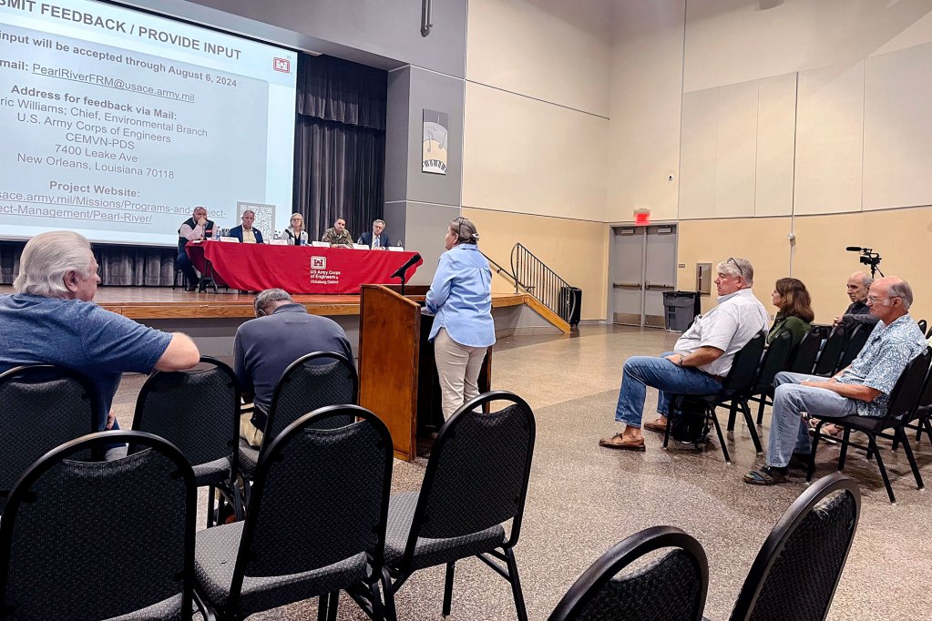 People at a public forum, one person standing at front to address the panel of seated people on the stage at the front of the room