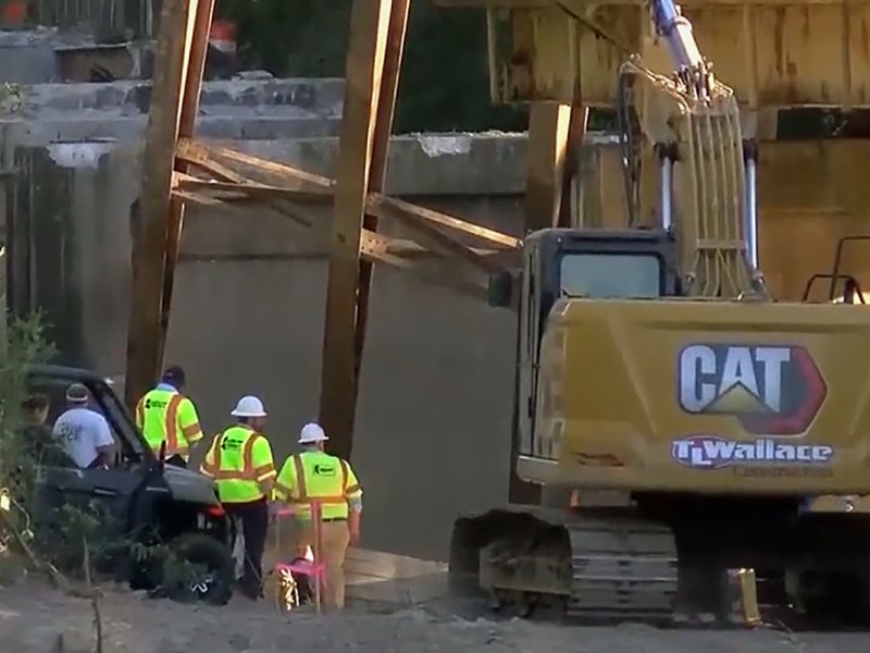 a screenshot shows construction workers and a CAT tractor at the site of a collapsed bridge