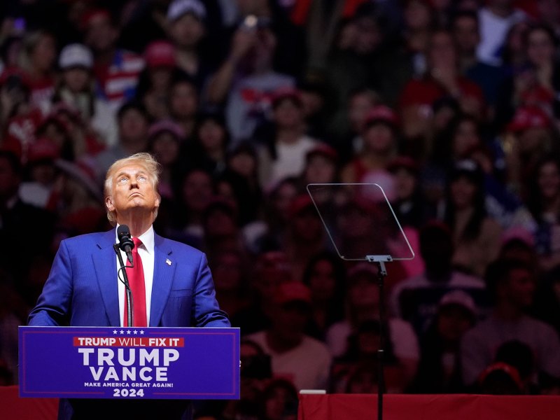 Donald Trump standing at a Trump/Vance podium with a large crowd visible behind him