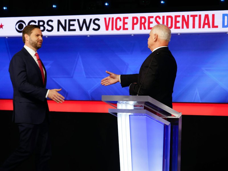 Two men go to shake hands behind a podium. A sign above them reads CBS News Vice Presidential Debate
