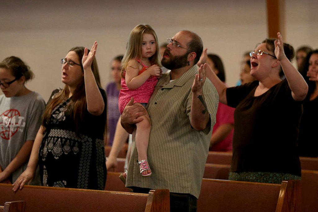 A man holds a toddler in a pink outfit as he and other people in church pews pray with their arms raised