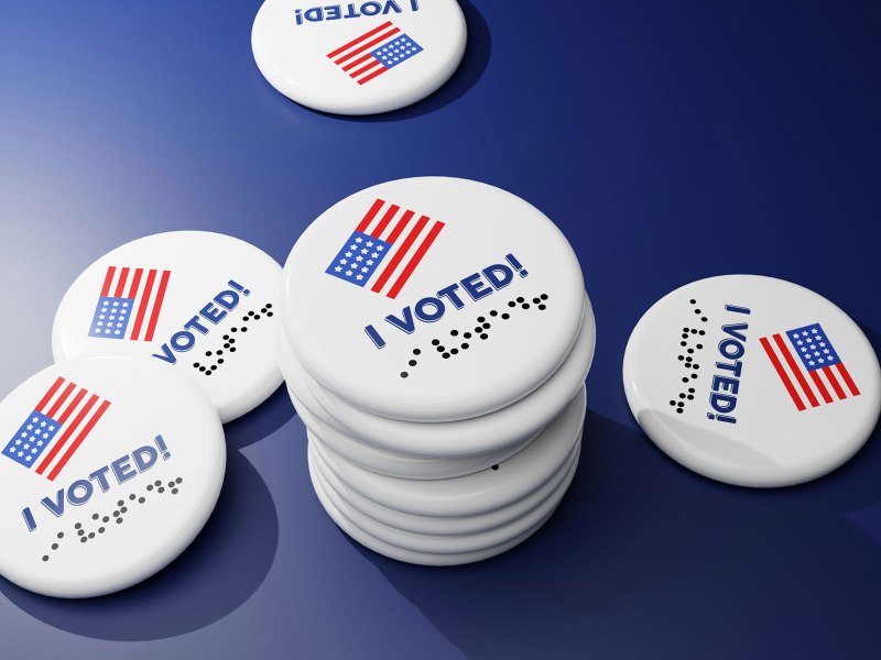 A stack of round white buttons that say I Voted, also in brail, with a US Flag on the top