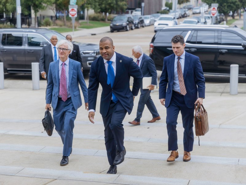 A Black man in a navy suit and blue tie walks up the steps of a courthouse with two White men wearing blue suits and holding leather briefcases.