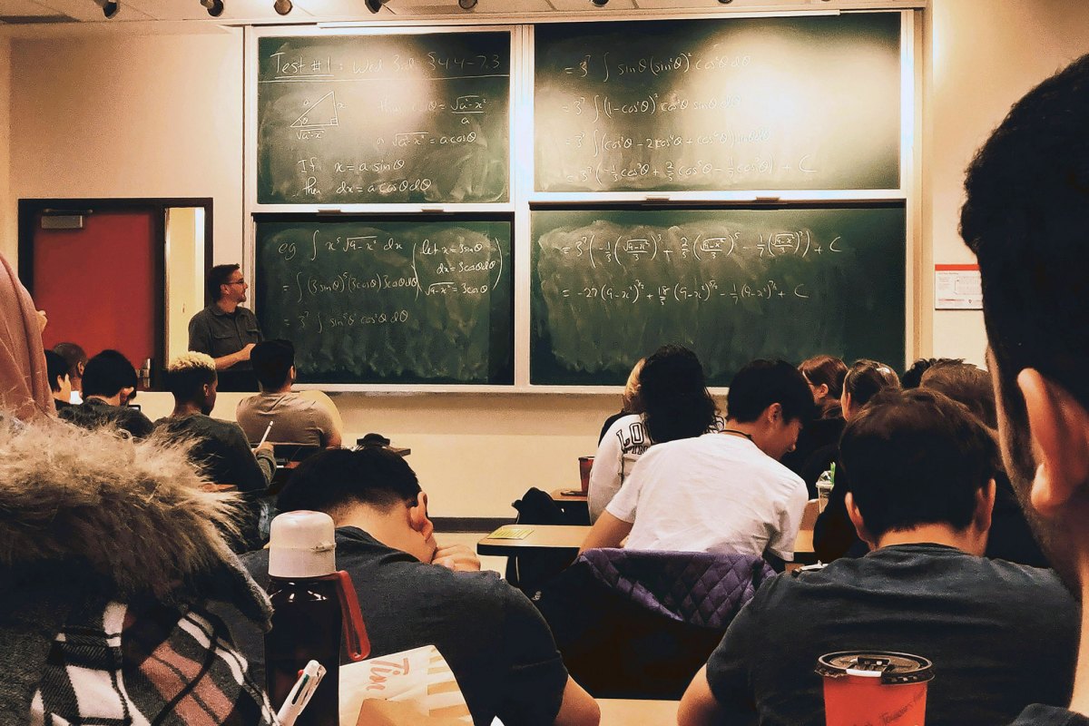 Students sitting in classroom desks in front of a chalkboard with mathematical equations written on it