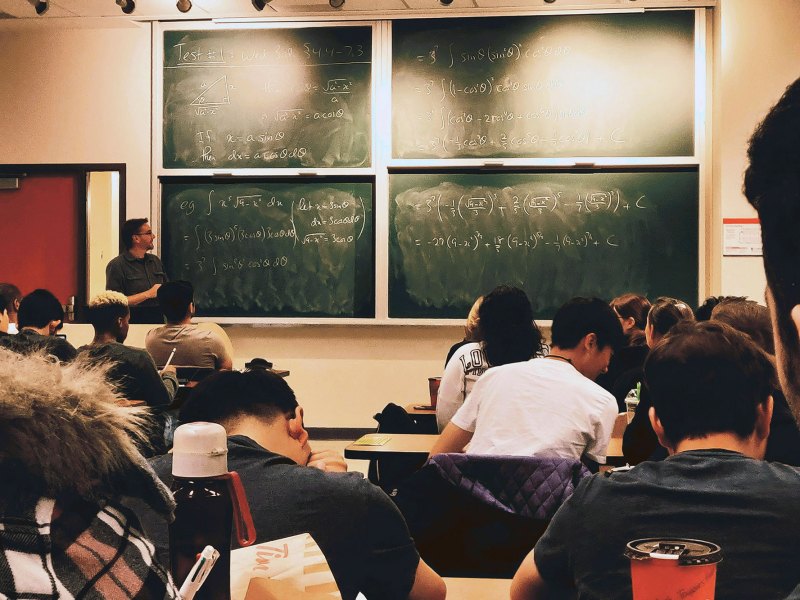 Students sitting in classroom desks in front of a chalkboard with mathematical equations written on it