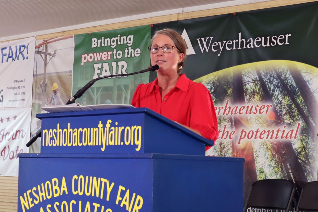 A woman in a red button down top and glasses speaks behind a bright blue podium that is labeled Neshoba County Fair