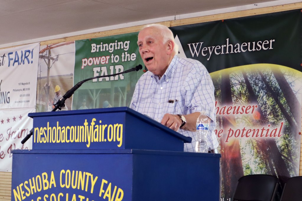 A man in a white button down top with light blue squares speaks behind a bright blue podium that is labeled Neshoba County Fair