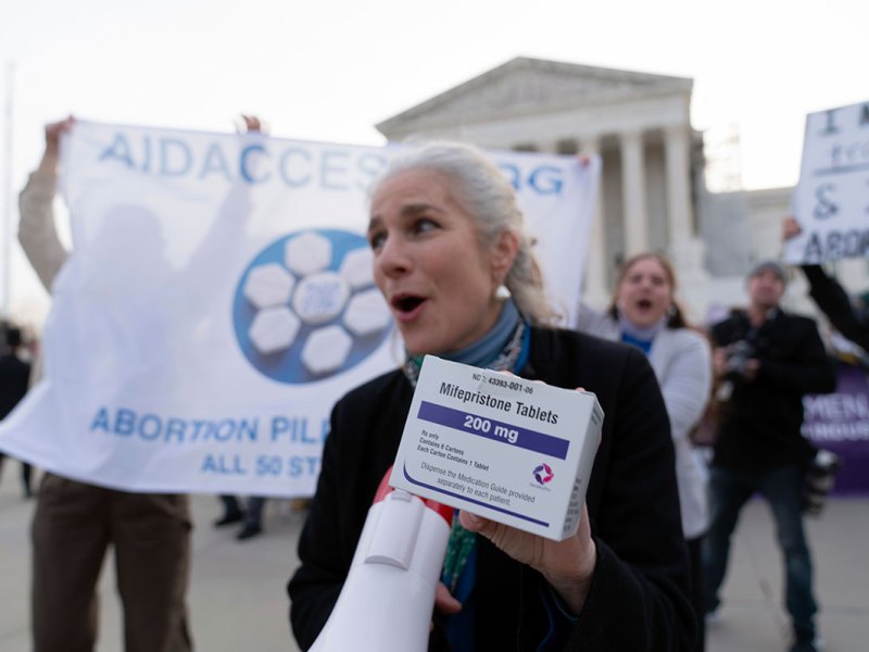 A person holds a box of mifepristone tablets in one hand and a megaphone in the other at an abortion rights rally