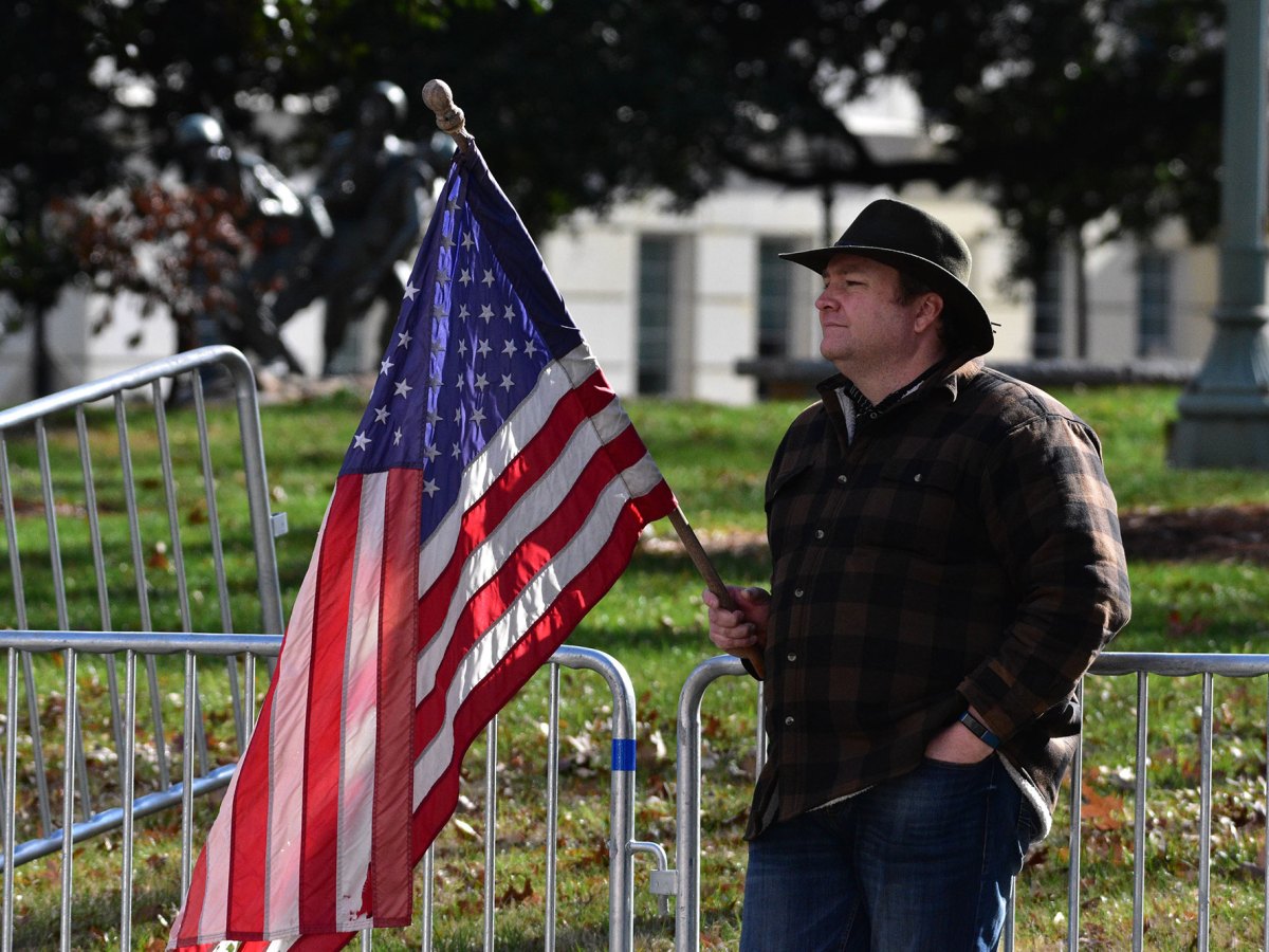 A man wearing a hat leans against a metal divider and holds a US flag