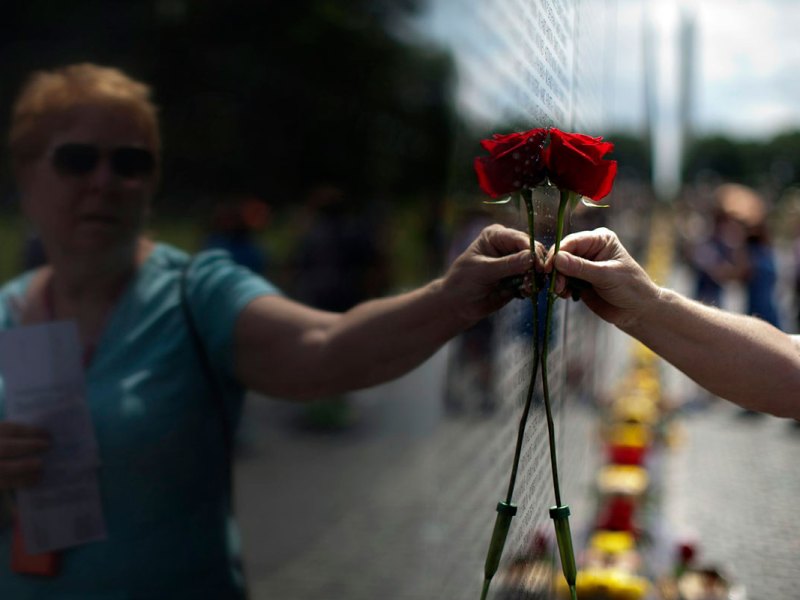 A woman holds a red rose up to a black wall inscribed with names