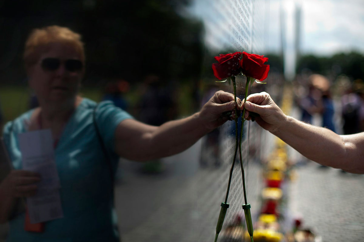 A woman holds a red rose up to a black wall inscribed with names