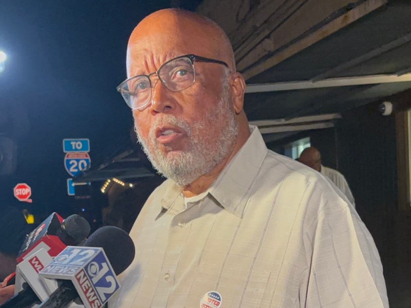 A bearded man wearing glasses speaks into reporter microphones outside at night