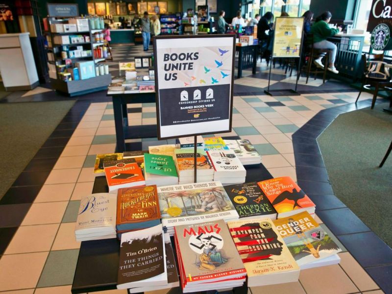A pile of books sits on a table in a book store, with the sign "Books Unite Us" above it. People sitting in the cafe can be seen in the background.