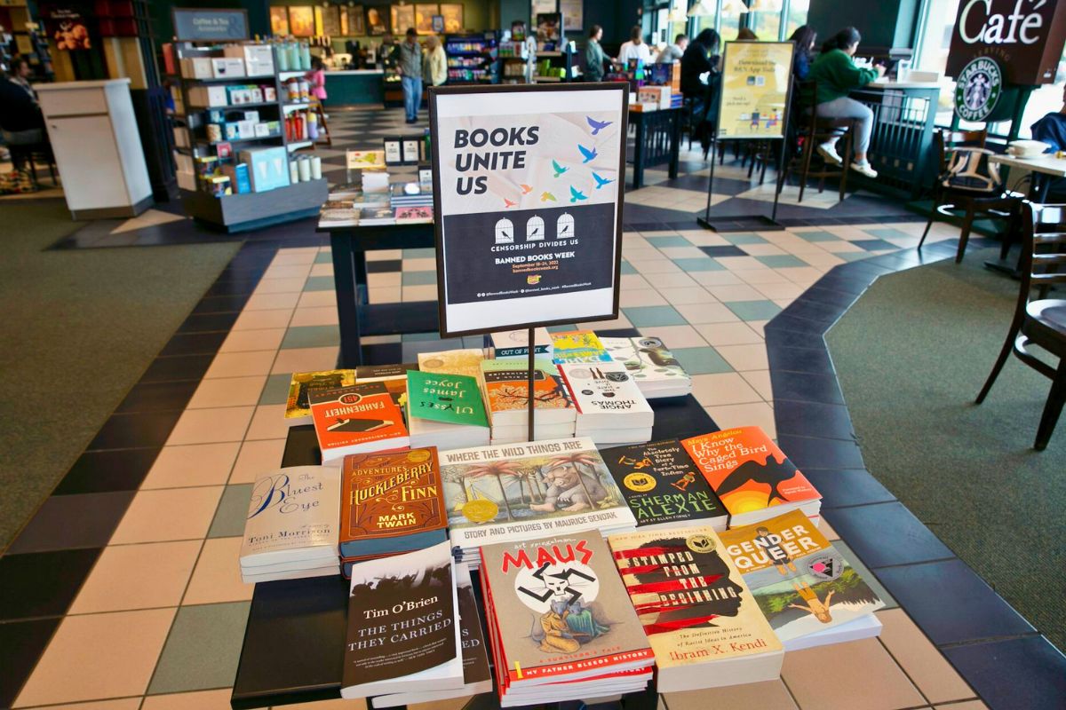 A pile of books sits on a table in a book store, with the sign "Books Unite Us" above it. People sitting in the cafe can be seen in the background.