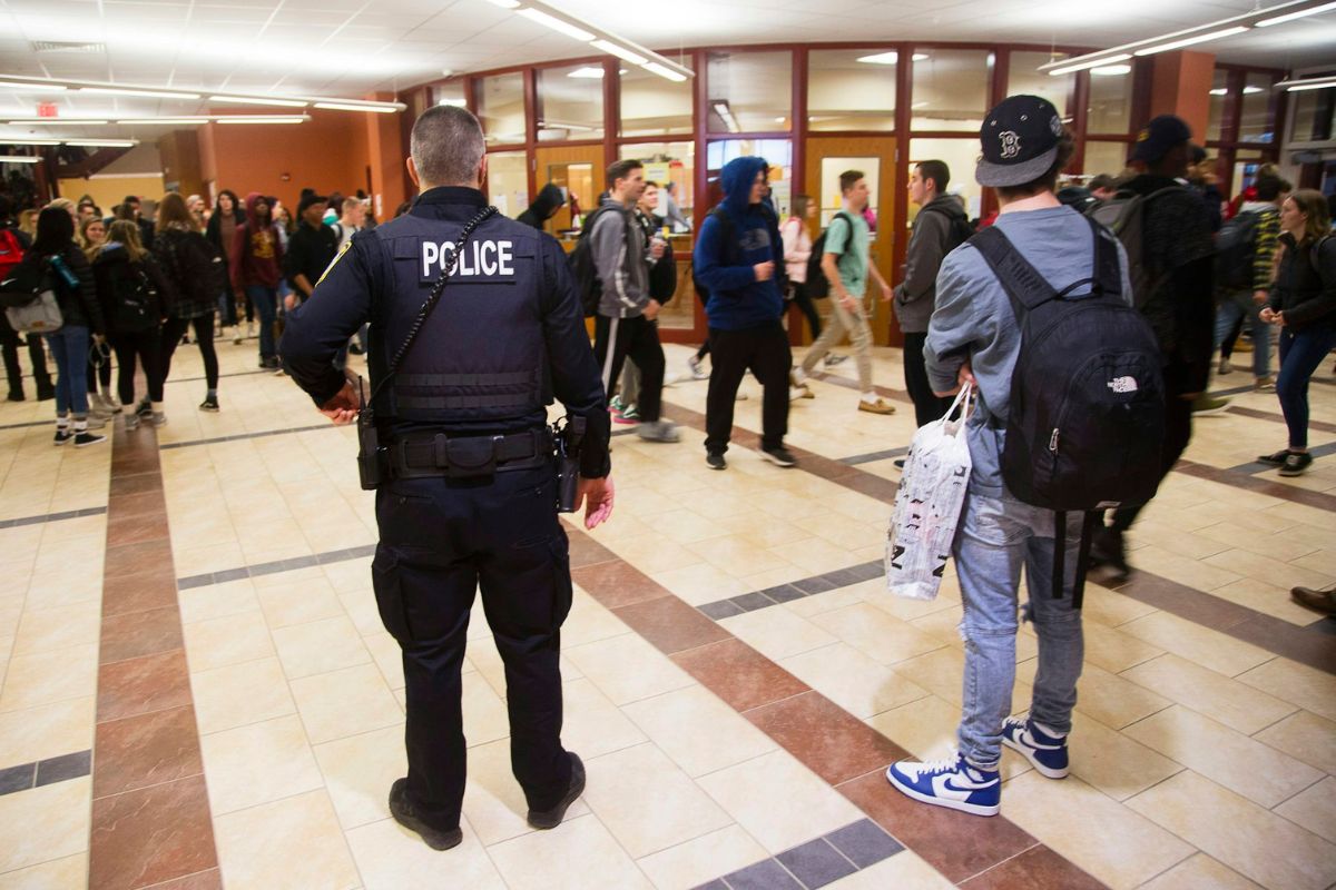 A person in full black Police gear stands in a high school hallway as teenagers walk through