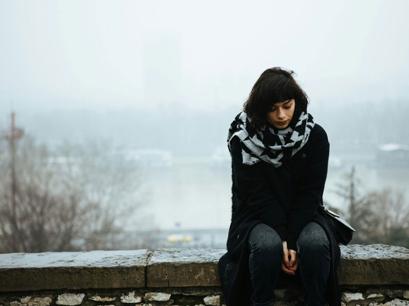 A woman in black sits on a stone wall outside during a foggy day