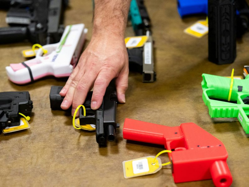 A hand reaches for a black 3D printed gun. Other guns are also on the table, some in bright colors