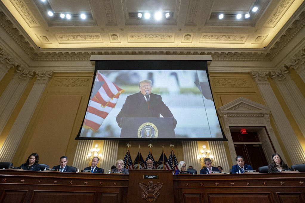 A row of men and women sit at a long wooded table with the US Seal on front. Above them is a projection showing Donald Trump