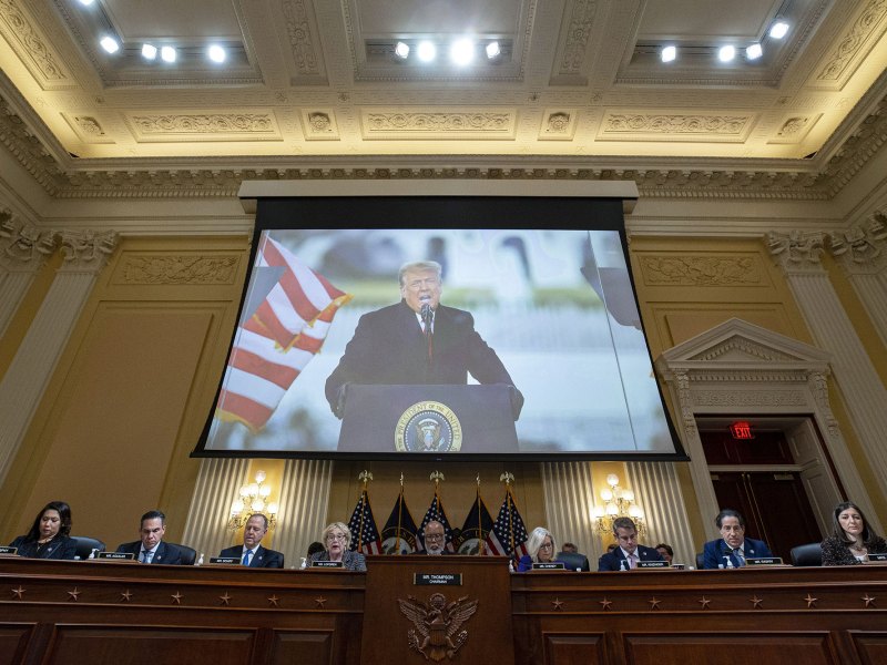 A row of men and women sit at a long wooded table with the US Seal on front. Above them is a projection showing Donald Trump