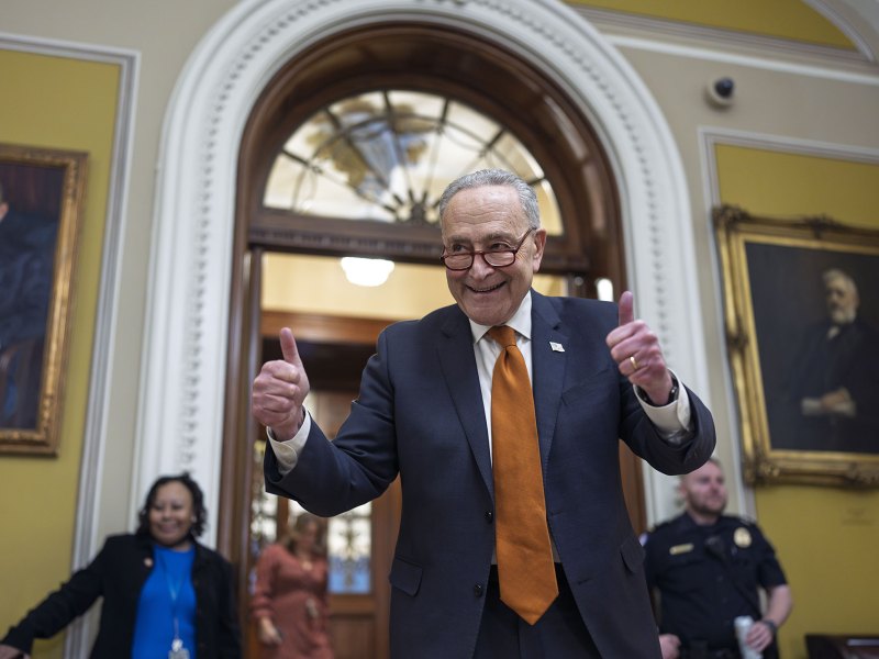 A older man in a suit and brown tie does a smiling pose with two thumbs up