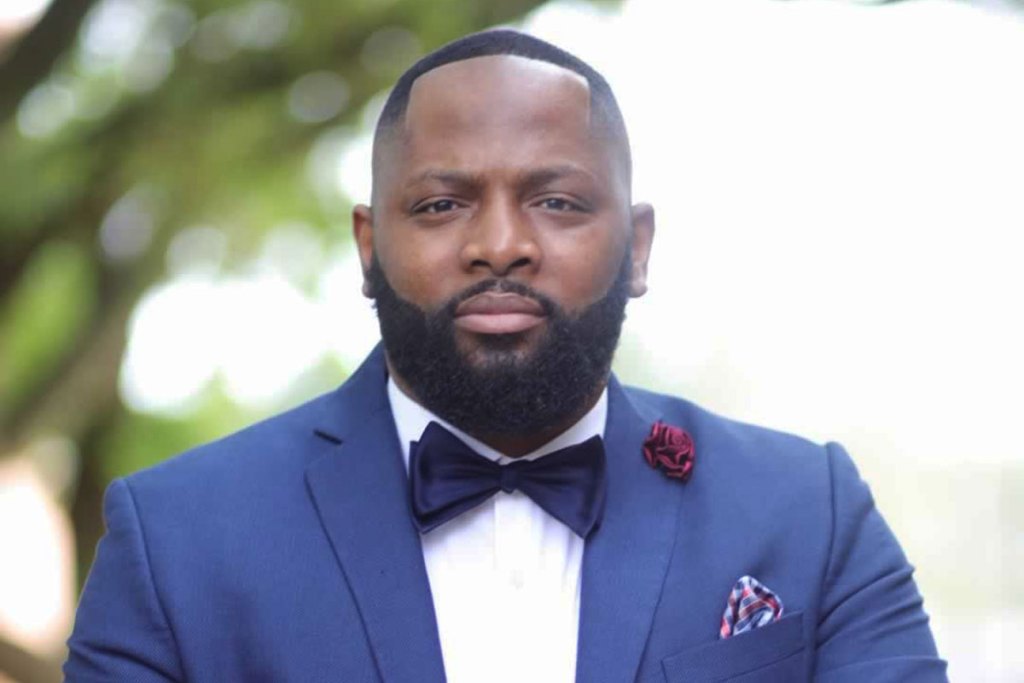 A close up of a bearded man in a blue suit and black bowtie standing outside