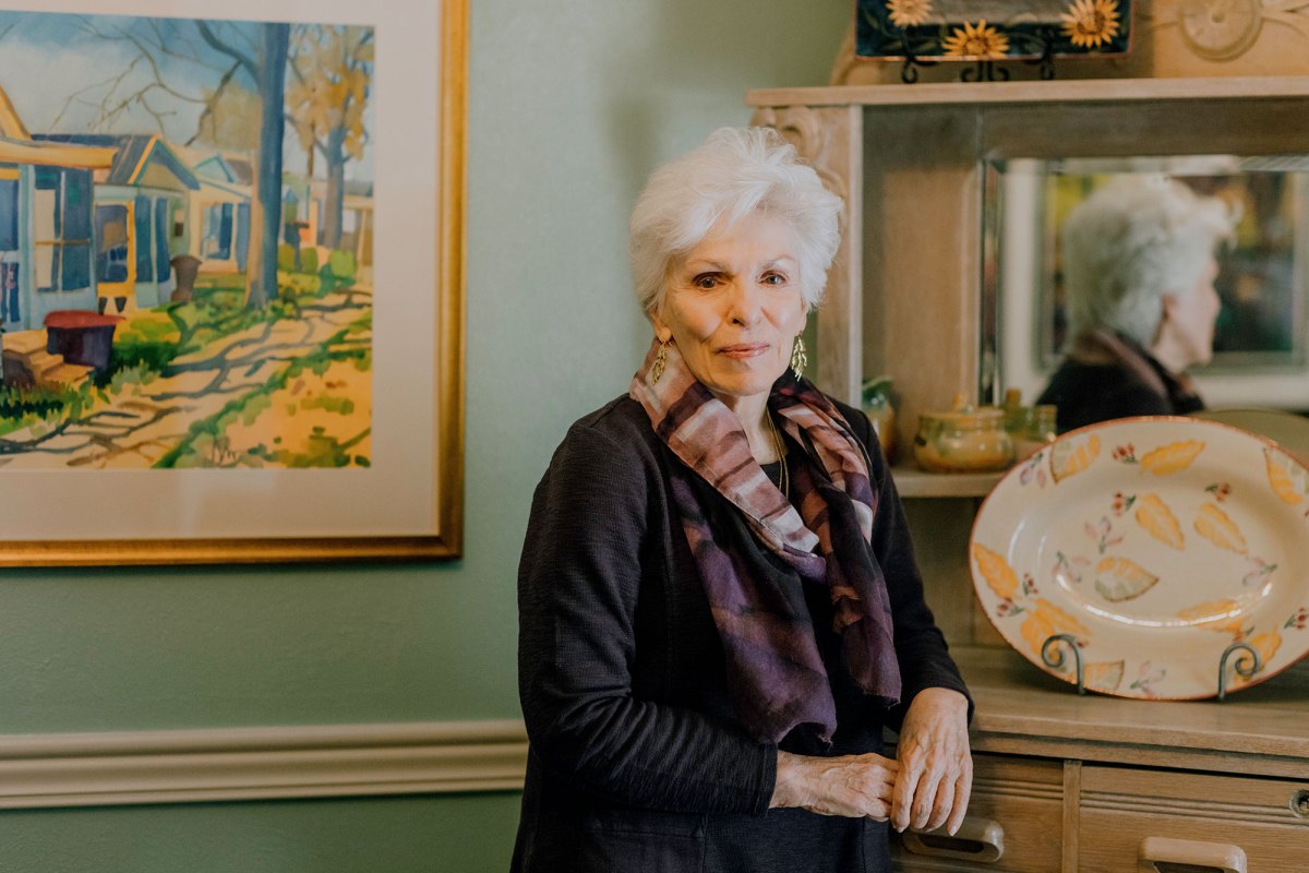 A woman in dark clothes and a patterned scarf poses beside a mirrored hutch inside a light green room