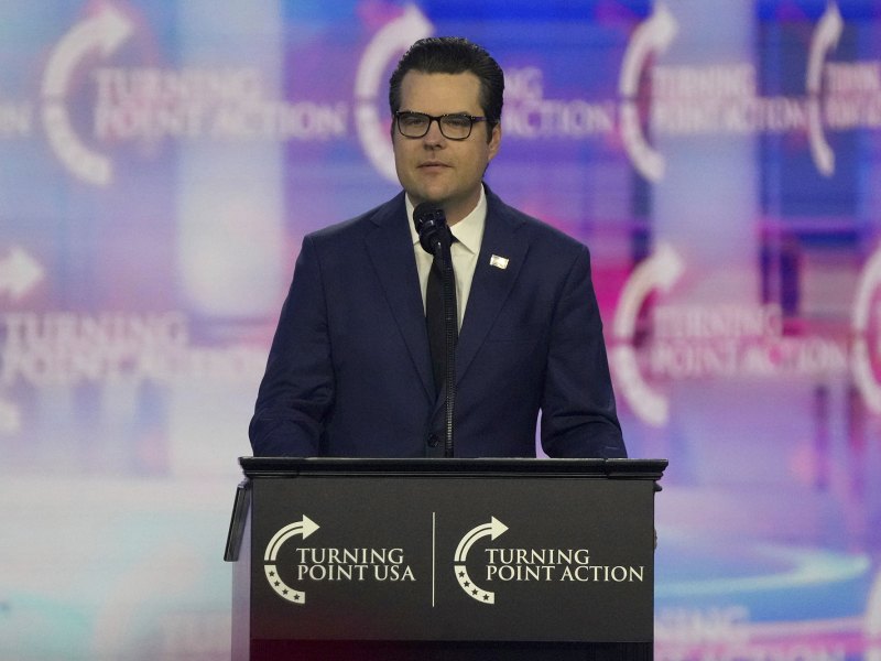 A man in a navy suit stands behind a podium on a stage. The logo for Turning Point USA is repeated everywhere.
