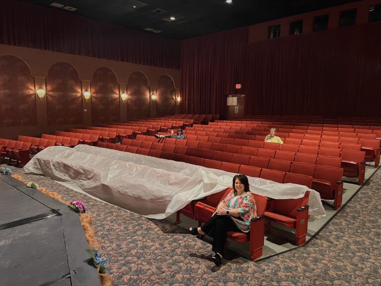 Three people sit spaced apart in red theater seating. The front two rows are mostly covered with a clear tarp
