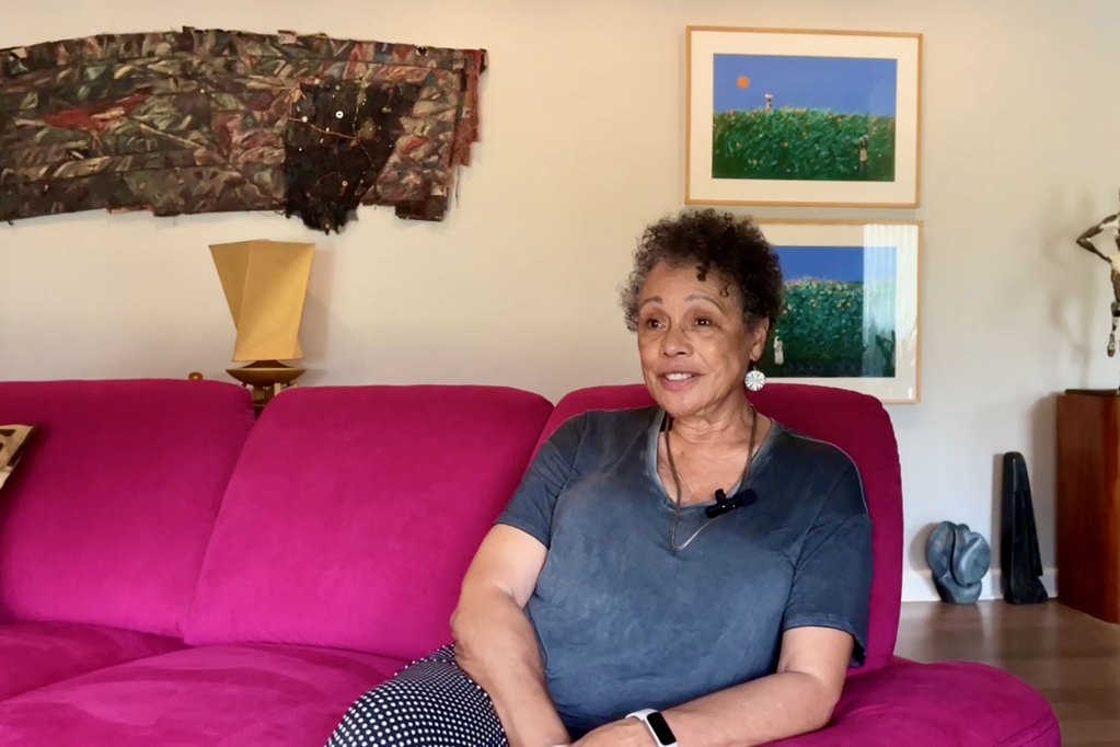 A woman seated in a living room on a bright fushia couch, art visible on the walls behind her