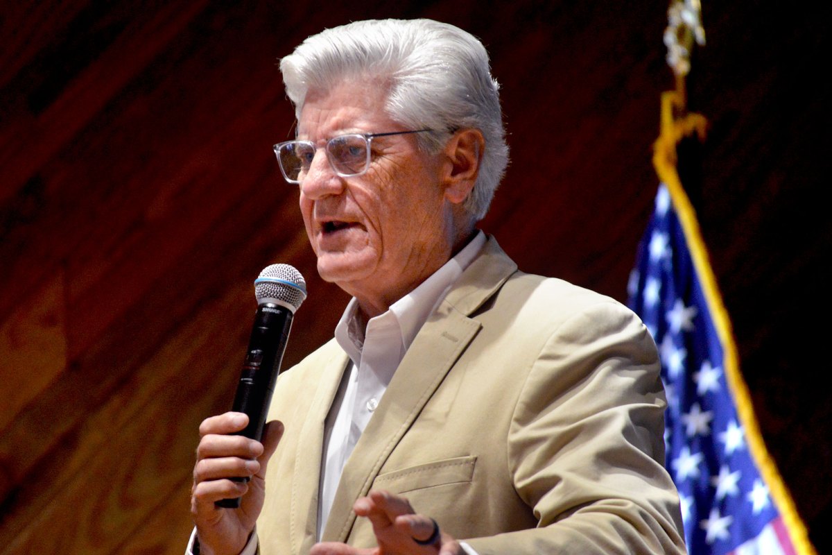 A man with white hair and glasses speaks on stage with a mic in hand