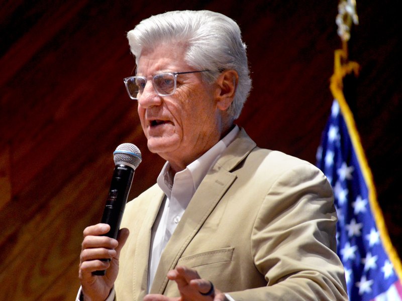 A man with white hair and glasses speaks on stage with a mic in hand