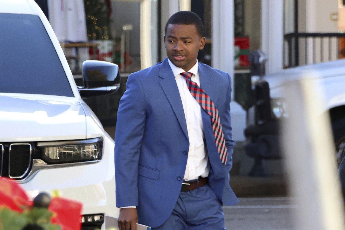 A man in a blue suit and red tie walks outside in front of a white vehicle