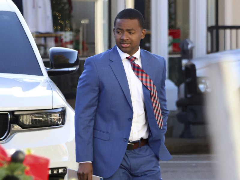 A man in a blue suit and red tie walks outside in front of a white vehicle