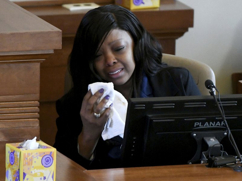 A woman sits an a court stand, crying with a tissue in hand