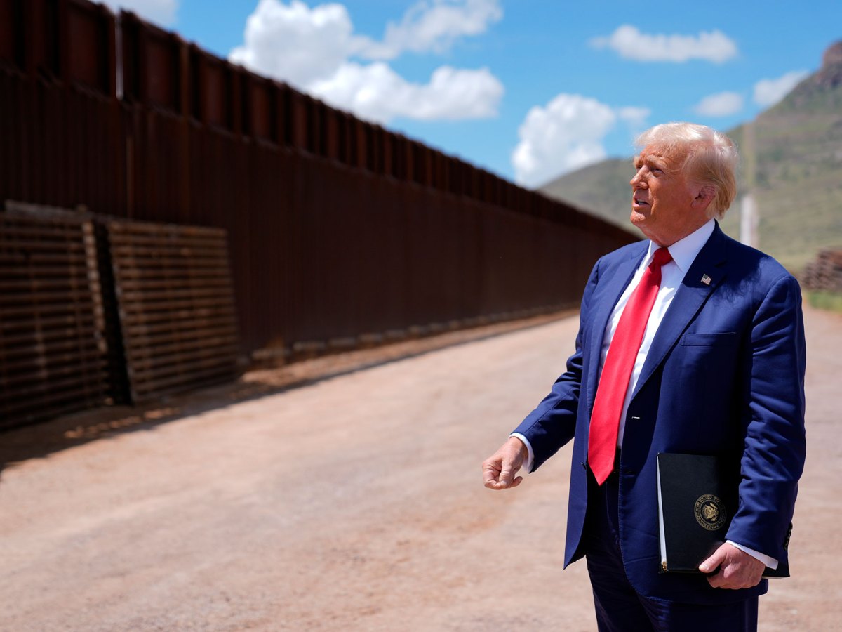 A older man in a blue suit and red tie looks up at a tall metal wall