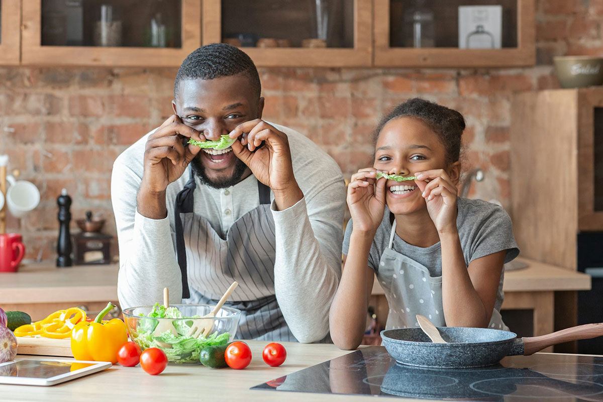 A father and child making silly faces in the kitchen, holding lettuce up as mustaches