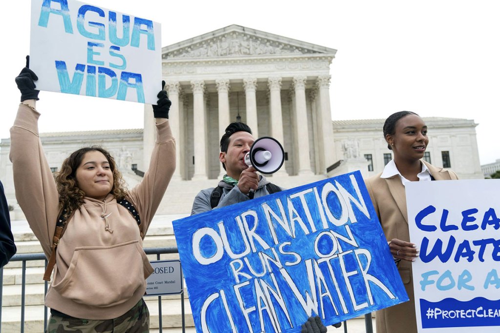 "People holding signs stand outside a marble building with pillars.