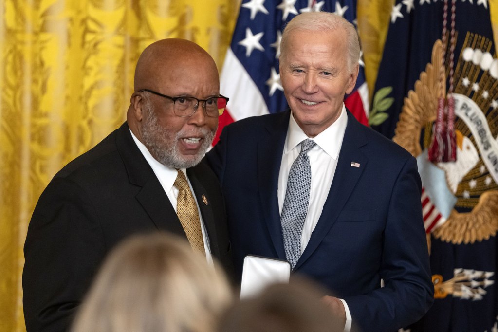 Two older men pose in front of a gold curtain and US Flags