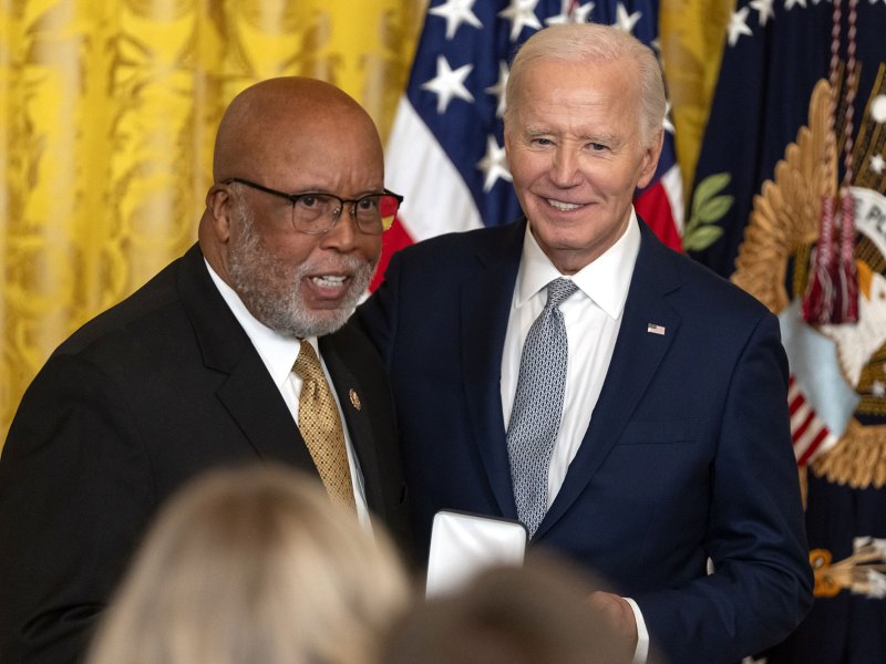 Two older men pose in front of a gold curtain and US Flags