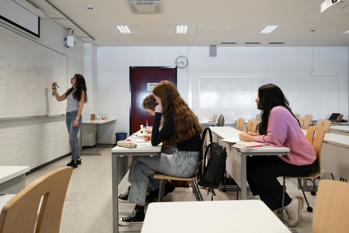 Students sitting at desks in a classroom as someone writes on the whiteboard