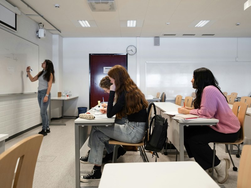 Students sitting at desks in a classroom as someone writes on the whiteboard