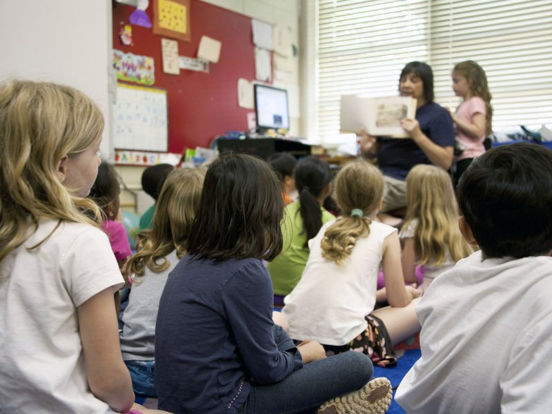 A teacher holds up a book to young students seated before her