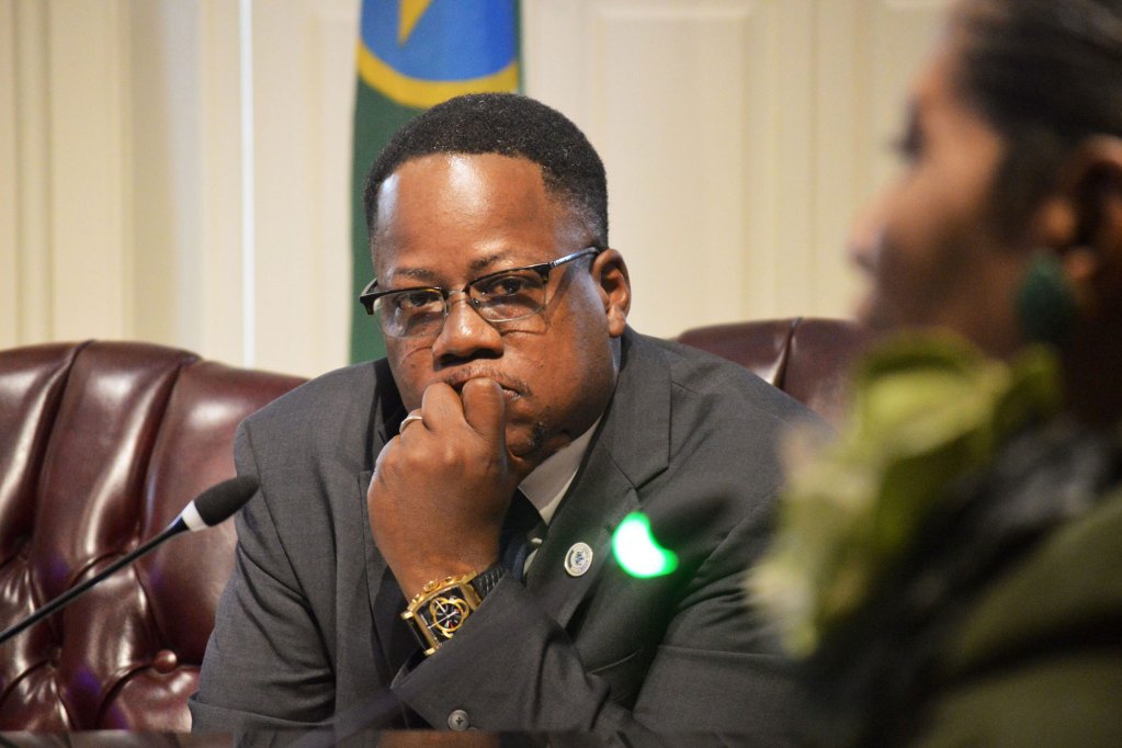 Black council member sitting inside of a council meeting.