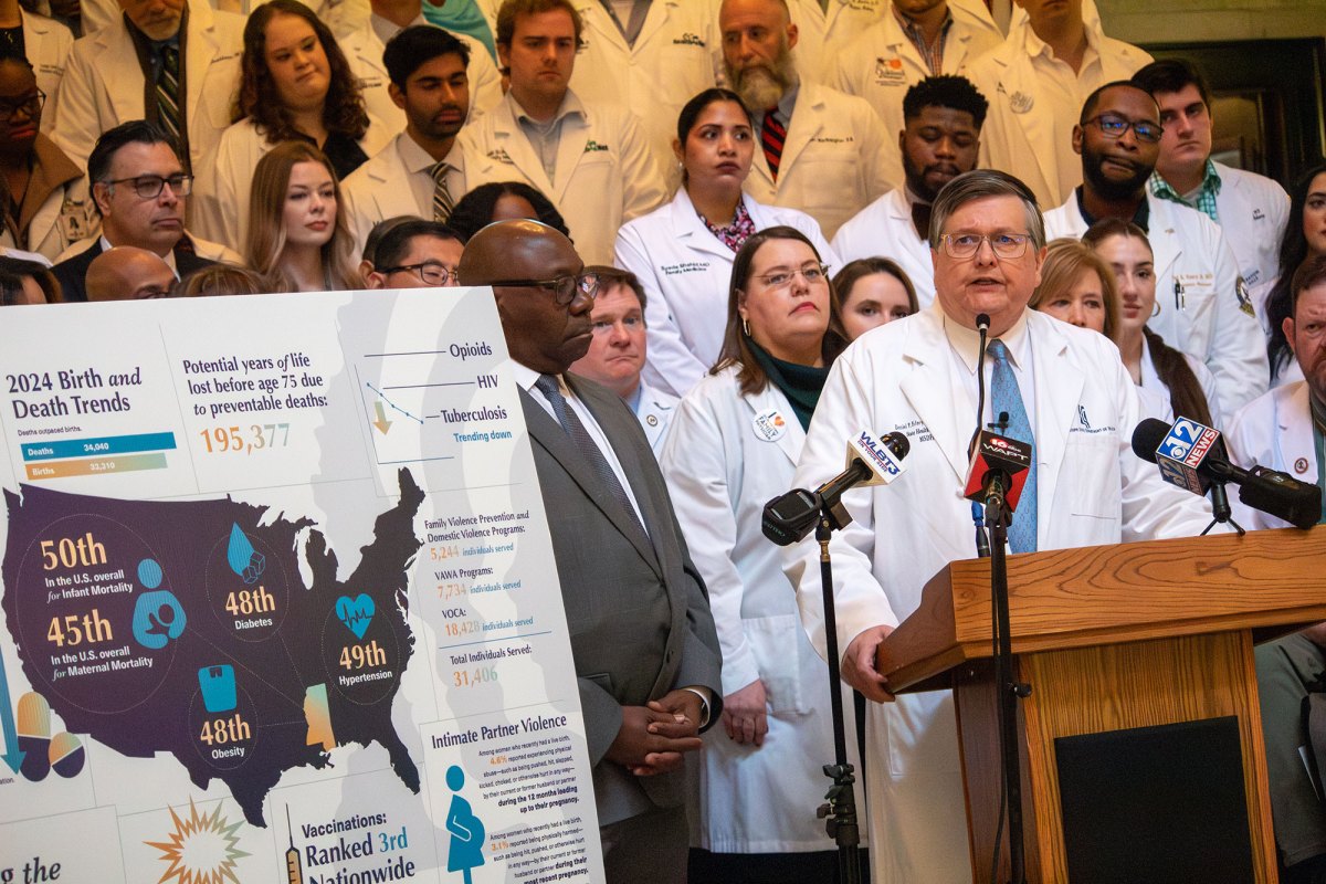 An older man in a doctor's coat speaks at a podium, many more people in doctor's coats behind him. A sign to the left reads 2024 Birth and Death Trends with markings all over a US Map